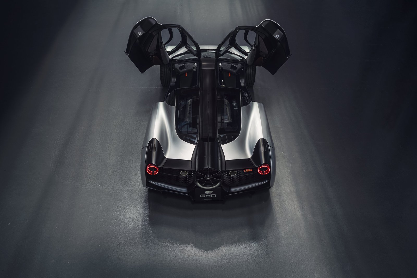 Overhead rear exterior shot of Gordon Murray T.50 silver sports car on dark surface, both upward-opening doors raised, revealing cockpit and rear vents.