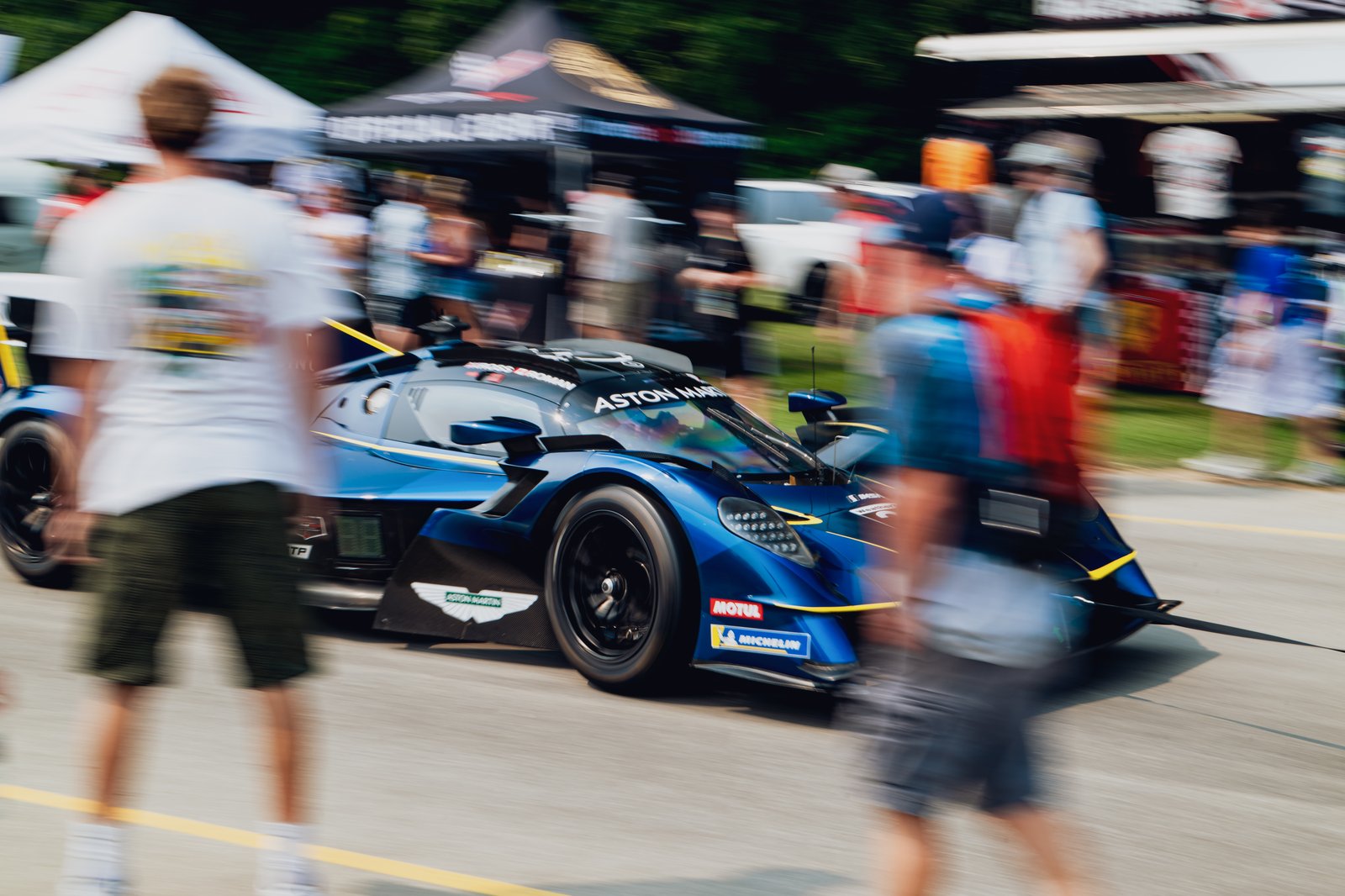 Aston Martin race car in motion, front 3/4 view at an outdoor event with spectators and tents in the background.