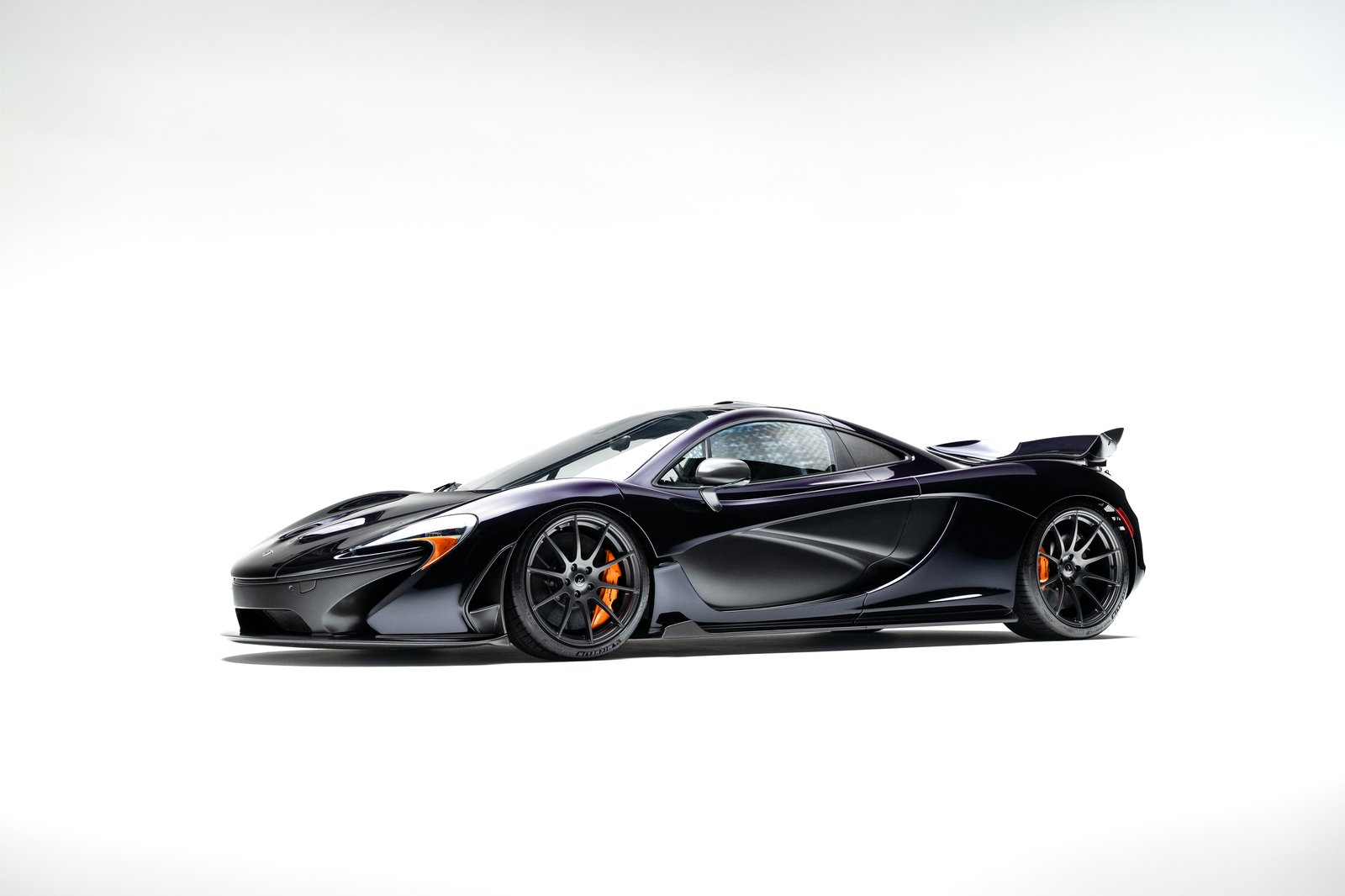 Side profile exterior shot of a 2015 McLaren P1 in dark finish on white studio background, with side intakes, raised rear wing, and orange calipers
