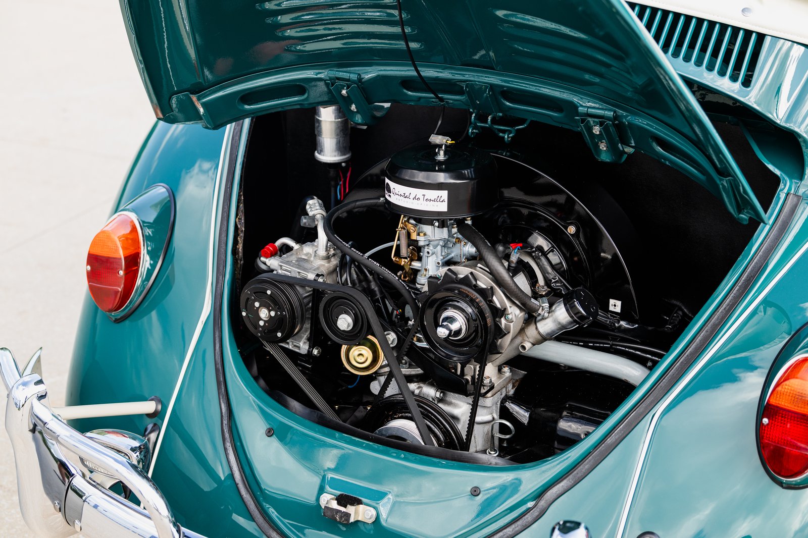 A teal 1964 Volkswagen Beetle is shown from the rear with the engine lid open, exposing the rear-mounted engine. The close framing includes chrome bumper details, both rear taillights near the edges, and vented decklid p