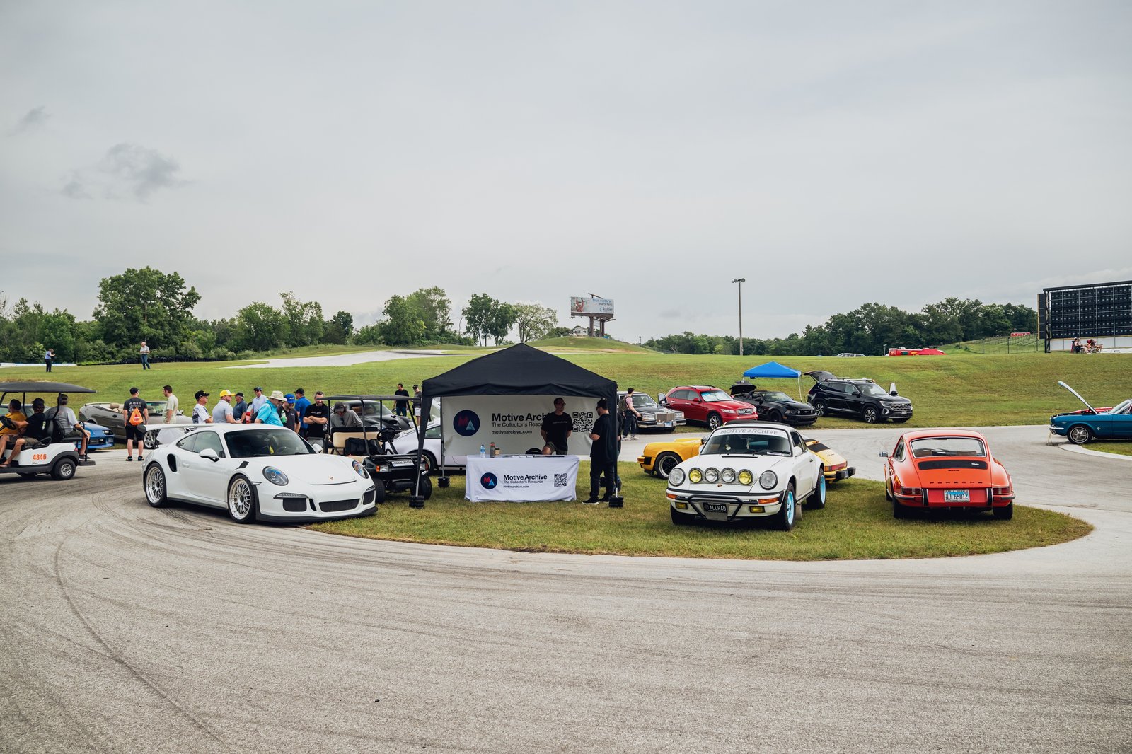 View of classic and modified cars at a car show with attendees near a promotional tent.