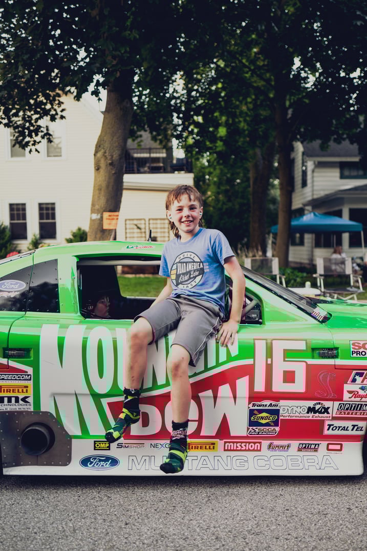 A green and red race car with sponsorship logos, a boy sitting beside it in an outdoor event setting.