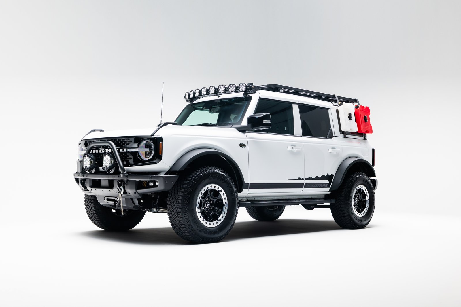A white four-door Ford Bronco Wildtrak is shown in a studio on a white background. The SUV has a roof rack with auxiliary lights, front bumper lighting and winch hardware, large off-road tires, black graphics, and side-mounted red and white containers.