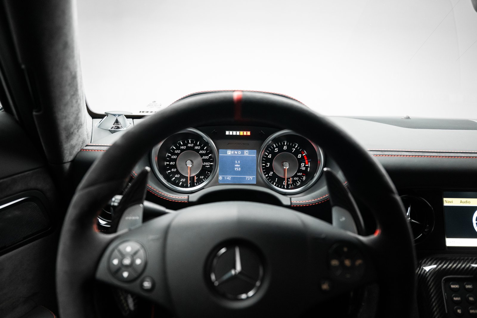 Interior view of a Mercedes-Benz SLS AMG focused on the steering wheel and instrument cluster. The dashboard shows black trim with red stitching, analog gauges, and a center infotainment screen partially visible on the right.