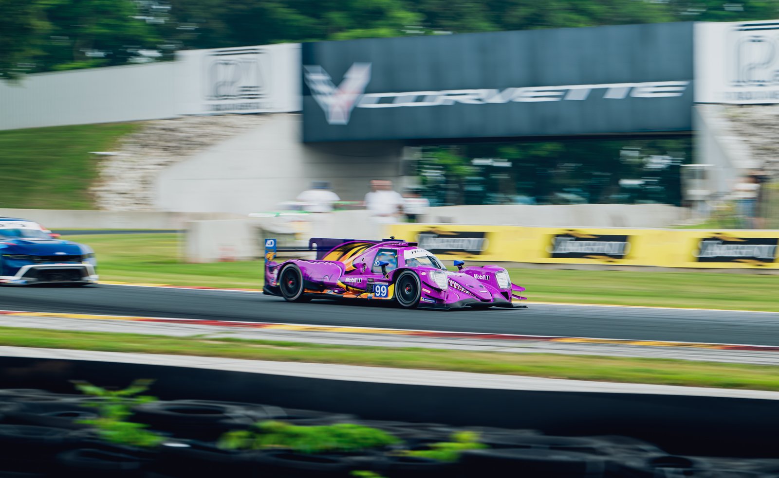 AO Racing's Spike LMP2 car, front 3/4 view in motion on a racetrack with blurred background of another car and banners.