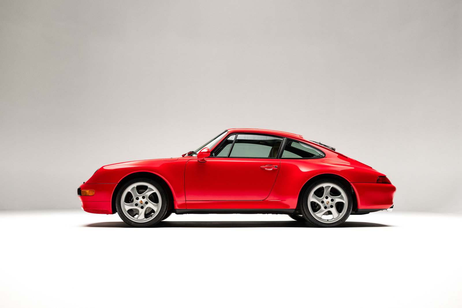 The image features a 1998 Porsche 911 Carrera S coupe in Guards Red, showcasing its sleek profile and distinctive design. The car is positioned against a neutral background, highlighting its body lines and 18-inch Techno