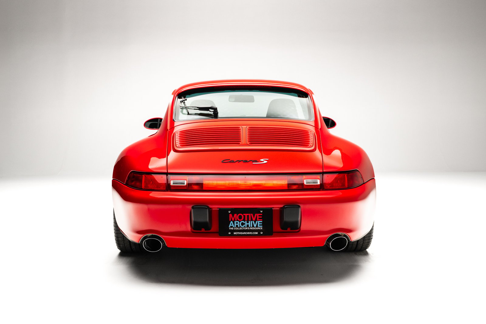 The image features a 1998 Porsche 911 Carrera S coupe in Guards Red, showcasing its rear view. Notable details include the split rear grille, dual exhaust outlets, and the 'Carrera S' badge on the rear.