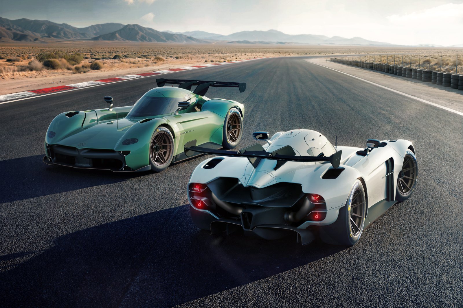 Two track-focused prototype race cars are parked on a wide desert racetrack with mountains in the background. The primary white car in the foreground is shown from the rear three-quarter view with illuminated taillights, exposed aero bodywork, and a large rear wing.
