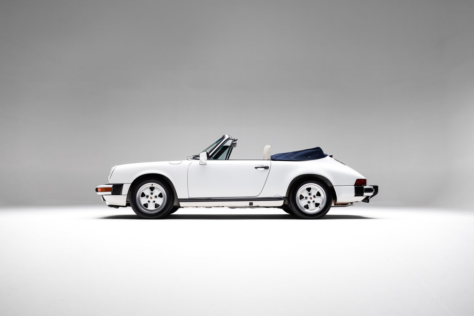 Full side-profile studio view of the 1988 Porsche 911 Carrera Cabriolet with the dark blue soft top folded.