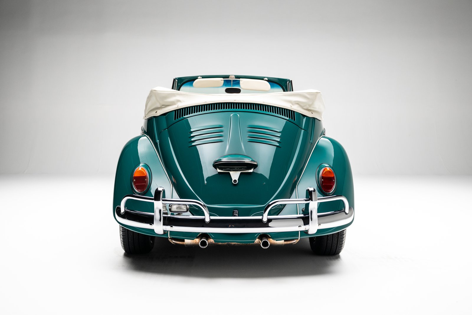 A teal 1964 Volkswagen Beetle convertible is shown from the rear in a white studio setting. The image highlights the chrome rear bumper, oval taillights, dual exhaust tips, vented engine lid, and folded white soft top.