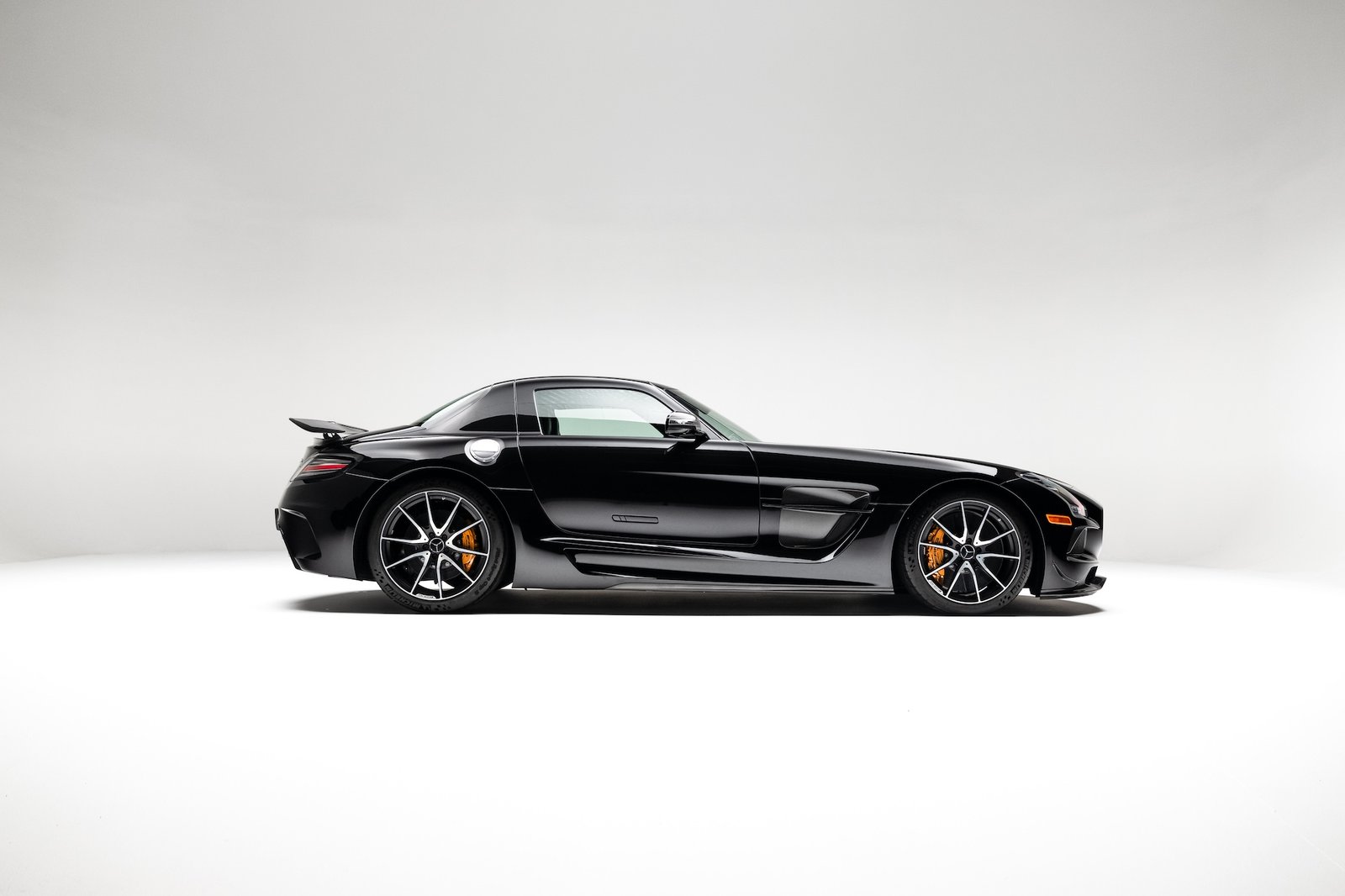 A black Mercedes-Benz SLS AMG coupe is shown in a studio on a seamless white background. The car is viewed in full side profile with a low stance, long hood, orange brake calipers, and a small rear spoiler visible.