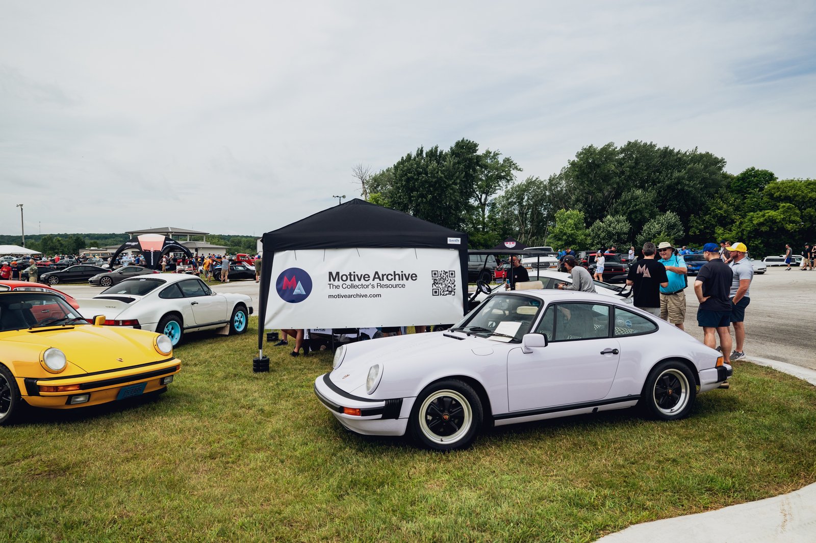 2025 Porsche 911 front 3/4 view at an outdoor car show with attendees and branding tent in the background.