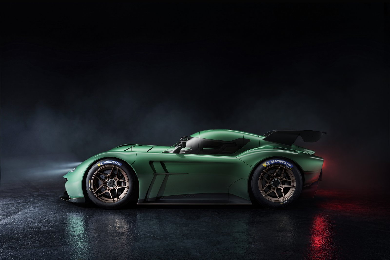 A green low-slung race car is shown in side profile on a dark studio floor with misty lighting. It has exposed Michelin racing tires, a large rear wing, and red rear lighting reflections on the wet surface.