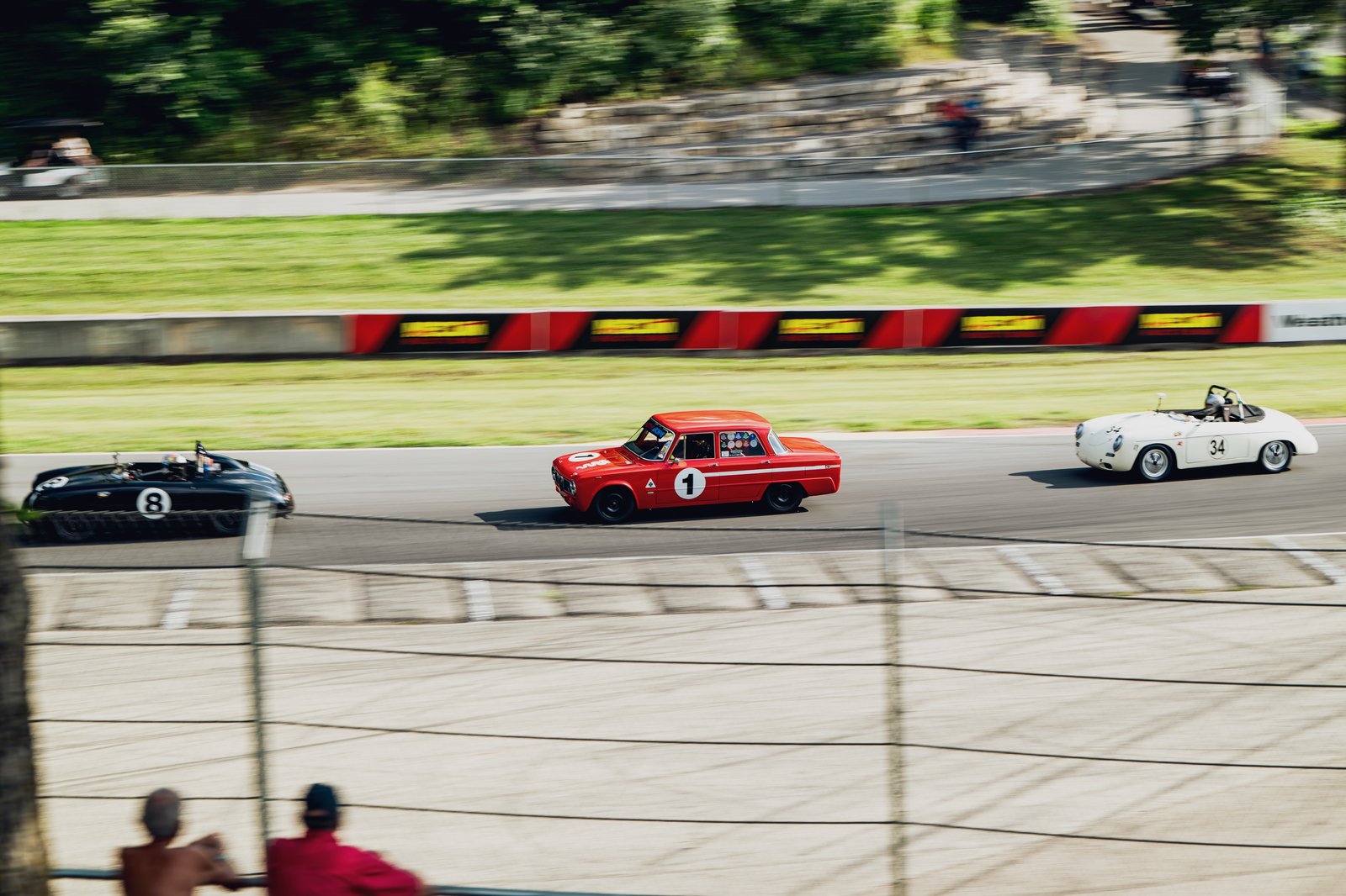 Three vintage race cars in motion on a racetrack, front 3/4 view, with a red car leading the pack.