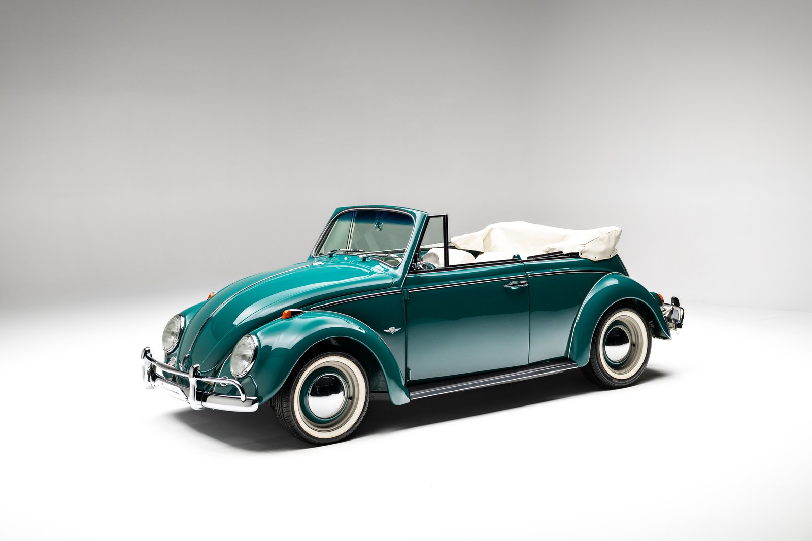 A teal 1964 Volkswagen Beetle convertible is shown in a studio on a white seamless background. The car has a folded white soft top, chrome bumpers, round headlights, and whitewall tires.
