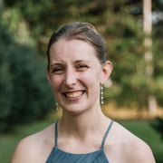 Smiling woman with shoulder-length hair, wearing a blue dress and dangling earrings. She's outdoors with a blurred green background, exuding a joyful and friendly demeanor.