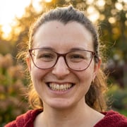 A warm, close-up candid photo of this beautiful woman smiling genuinely at the camera, outdoors in the golden hour.
