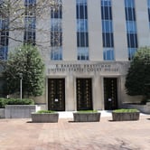 Two trees and several small bushes near the entrance to a multi-story building, with "E. Barrett Prettyman United States Courthouse" in black lettering above three tall doors.
