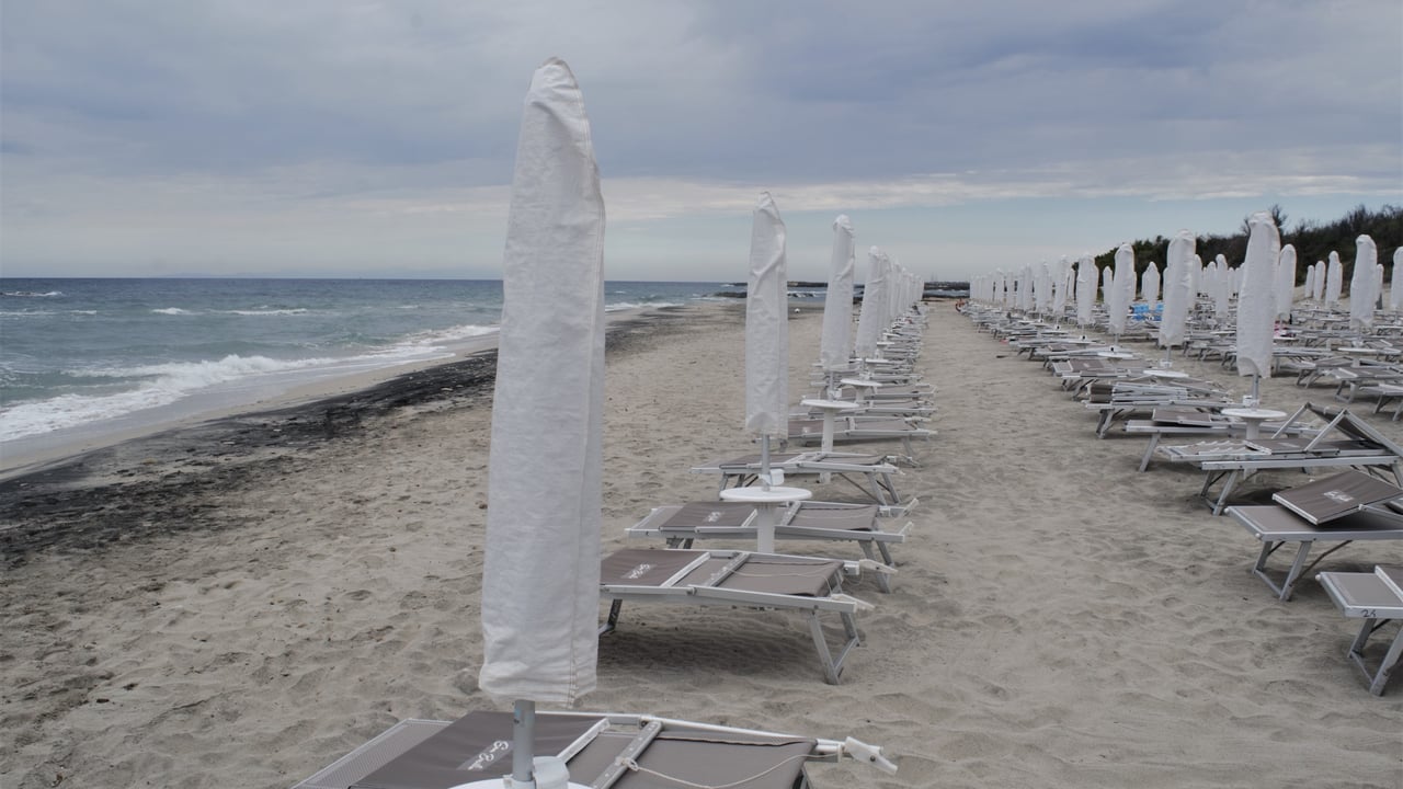 Sun beds and closed umbrellas on a beach under cloudy skies.