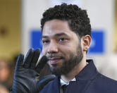 Wearing a black glove, Jussie Smollett waves with his right hand while smiling.