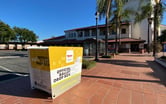 A amber colored ballot drop off box sits in the periphery of a white and adobe colored Spanish Colonial Revival building in Orange County.