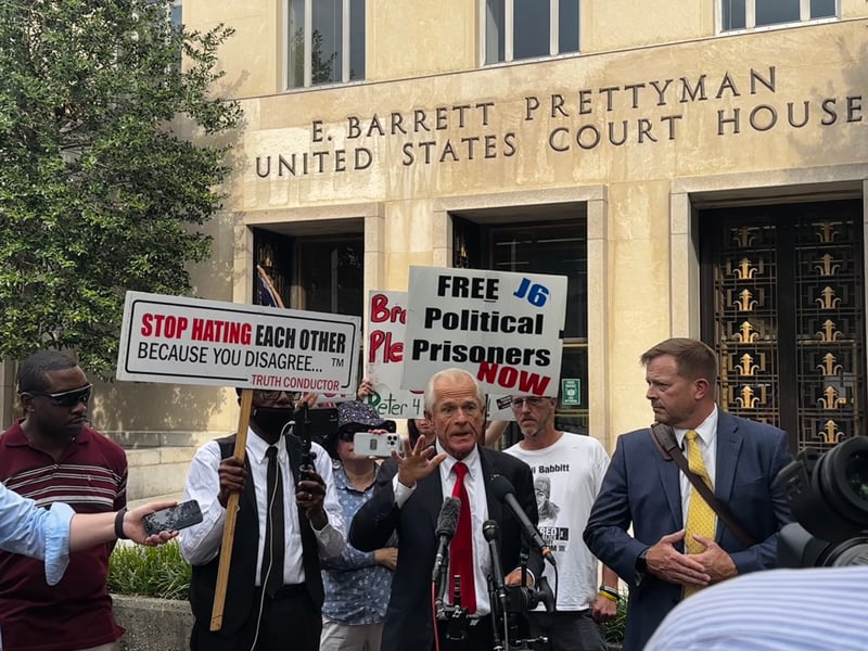 Peter Navarro speaking outside the D.C. federal courthouse with protestors holding supportive and critical signs behind him.