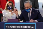 House Speaker Nancy Pelosi and Senate Majority Leader Chuck Schumer are signing a document sitting on a table, both are wearing masks, a banner at the bottom of the image says in all-caps "The American Rescue Plan"
