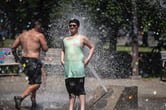 A man wearing a tank top gets sprayed by a water fountain, as a shirtless man runs toward it.