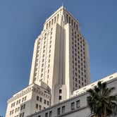 View of the tall, white Los Angeles City Hall building against a clear blue sky, with a palm tree in the foreground.