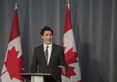Justin Trudeau gestures while speaking from a podium, with two Canadian flags behind him.
