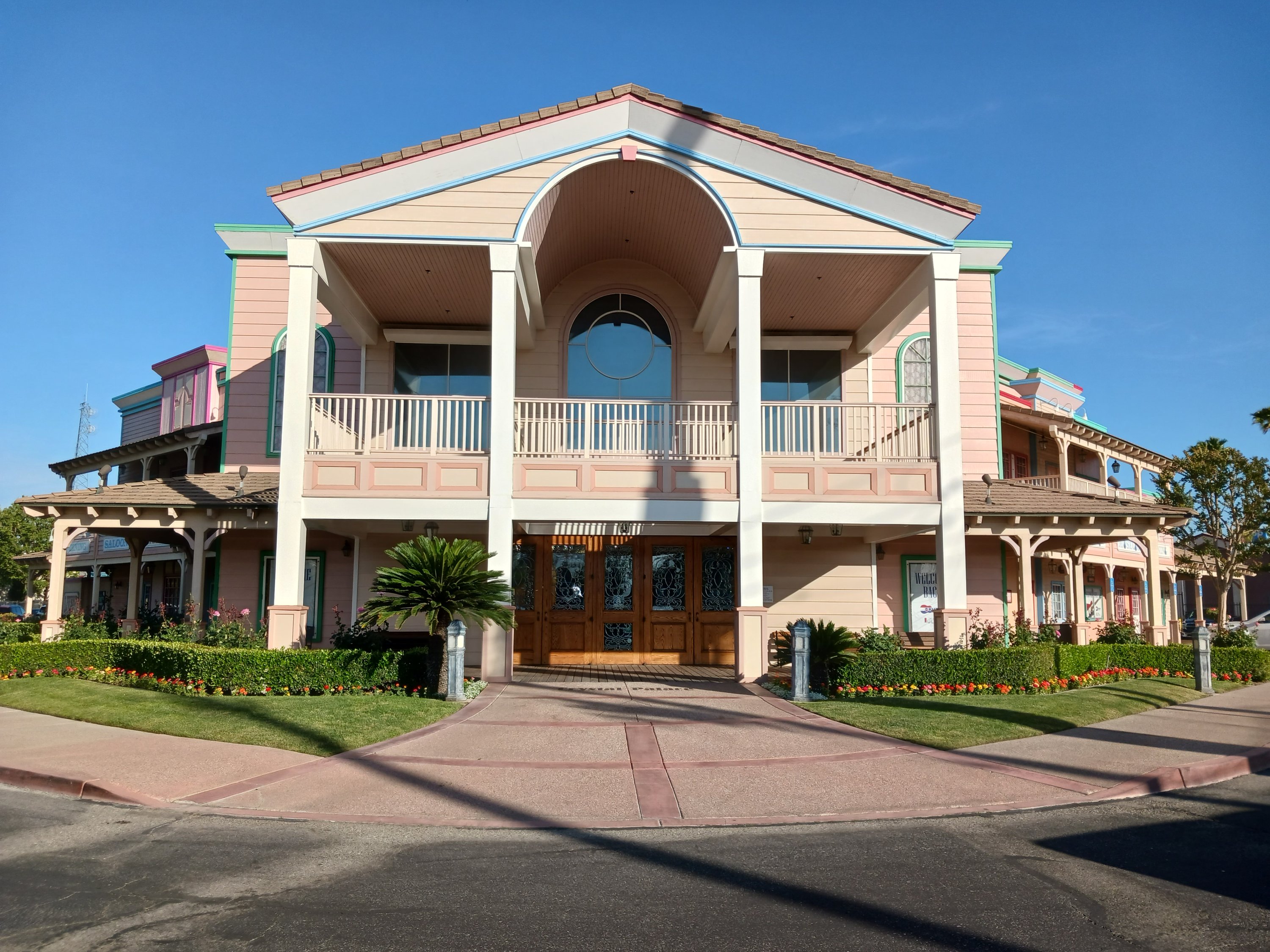 A wooden two-story building in the Western Revival style is bathed in afternoon sunlight. The building is pink and white with light blue trim and features elegant landscaping.