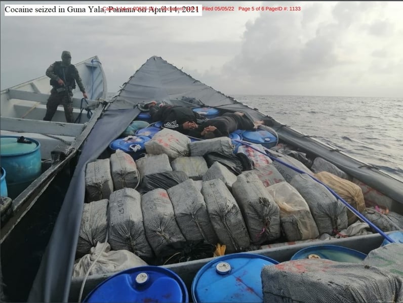 packages of cocaine are in a boat with two people lying on their stomachs with their hands behind their heads, a man in another boat holds a rifle