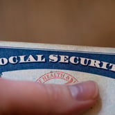 A person holds a U.S. Social Security card, with their thumb below "social security" in white letters at the top.