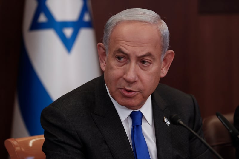 Benjamin Netanyahu sits while speaking during a meeting, with the Israeli flag in the background.