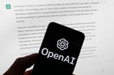 The OpenAI logo is seen on a mobile phone in front of a computer screen displaying text.