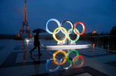 A man walks past the Olympic rings as it rains in Paris. The Eiffel Tower is in the background.