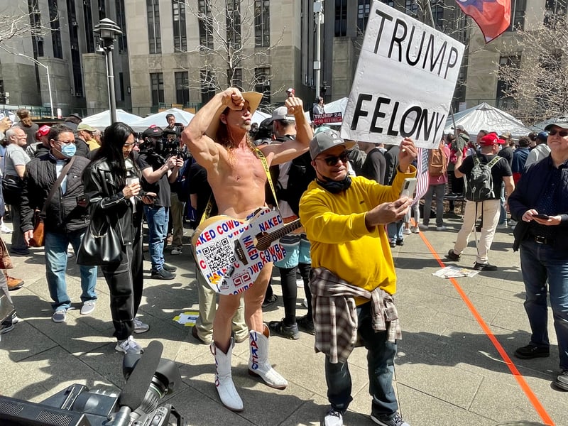a man in a cowboy hat poses flexing his muscles wearing a guitar that says "naked cowboy" and white boots, no other clothing is visible. a man next to him wearing a yellow sweatshirt and gray hat is holding a sign that says "Trump Felony" and is taking a selfie