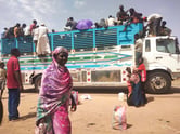 More than two dozen Sudanese people board a tall truck on a sunny day.