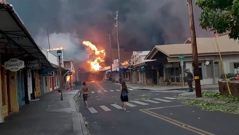 a photograph shows  two people on the street with their backs turned as they watch fires blazing in the background in Lahaina Town in Maui, Hawaii 