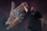 Killer Mike raises his right arm while holding a microphone in his left hand behind his back while performing on stage.