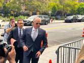 Silver-haired retired FBI New York office counterintelligence official Charles McGonigal arrives to the sidewalk entrance of the Manhattan federal court on a sunny morning wearing a blue suit, silver tie, and gold-rimmed aviator sunglasses. Photographers take pictures of him from behind and beside him as he enters, alongside his lawyer.