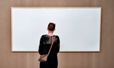 A person wearing a purse looks at an empty picture frame titled "Take the Money and Run" at an art museum.