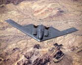 A black B-2 Spirit Stealth Bomber flies over a brown desert.