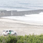 A white U.S. Border Patrol truck, with a green stripe across the center, parked on the beach, with the U.S.-Mexico border in the background.