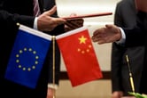 A person hands another person a folder, with miniature European Union and Chinese flags in front of them.