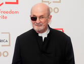 Salman Rushdie poses for photos while wearing a black glasses frame that covers his right eye.