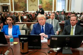 Donald Trump sits at the defense table in a court room wearing a blue suit and blue tie. His two attorneys, one male and one female, sit on either side of him.