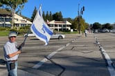 A white man in his 60s is waving the flag of Israel at a road intersection.