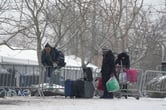 People walk near metal barricades with suitcases in the snow.