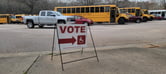 A red-and-white sign on a sidewalk that says "vote", with an arrow pointing to the right and a handicapped symbol . Six yellow school buses, a white bus, a silver truck, and three red sedans are in the parking lot behind the sidewalk. Bare trees are seen behind the vehicles.